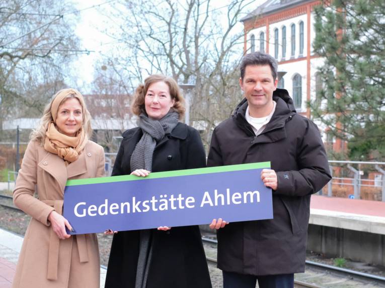 Drei Personen stehen an einer Haltestelle und halten ein Schild mit dem Namen der Haltestelle in den Händen.