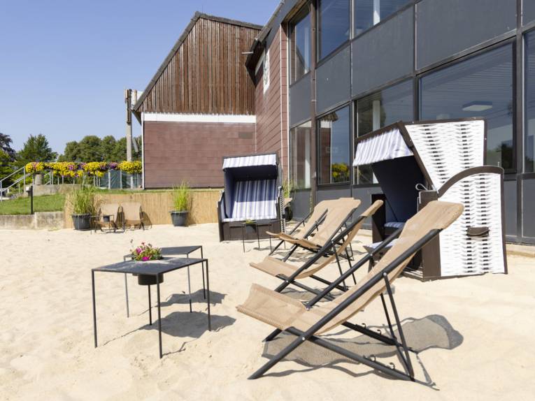 Liegest&uuml;hle, Strandk&ouml;rbe, Tische auf einer Sandfl&auml;che vor einem Geb&auml;ude stehend.