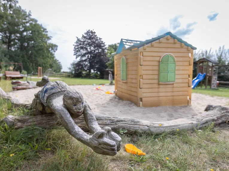 Im Vordergrund geschnitzter Schwimmer, Sandkiste mit Spielhaus