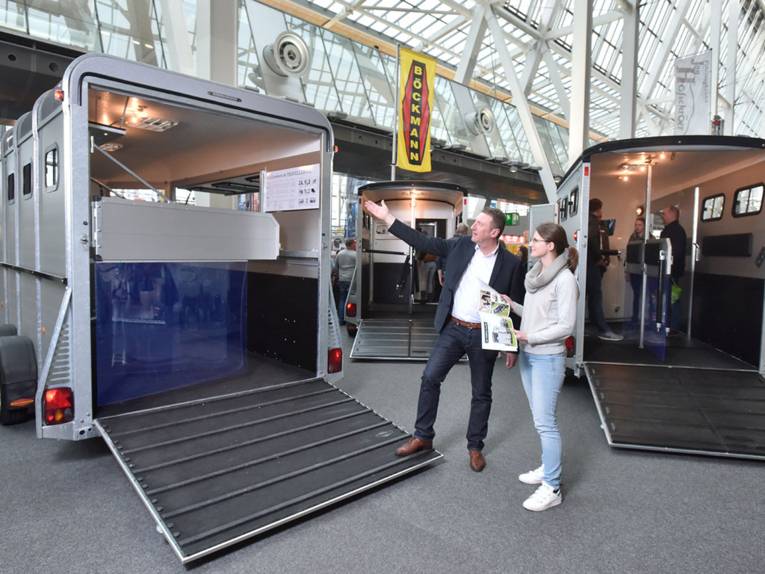 Ein Mann präsentiert einer Frau einen geöffneten Pferdeanhänger.