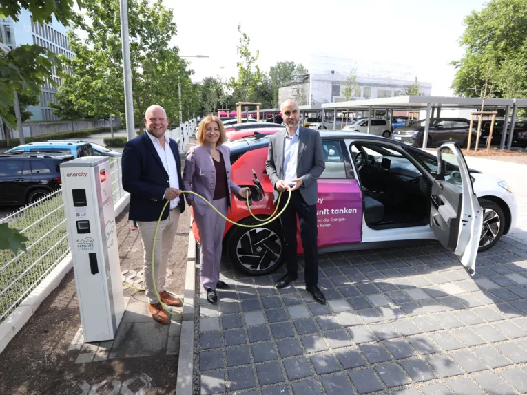 Zwei Männer und eine Frau stehen vor einem Auto an einer E-Ladesäule.