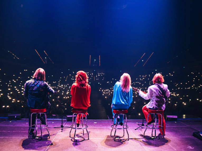 Zwei Männer und zwei Frauen sitzen mit Mikrofonen und Musikinstrumenten auf Barhockern auf einer Bühne.