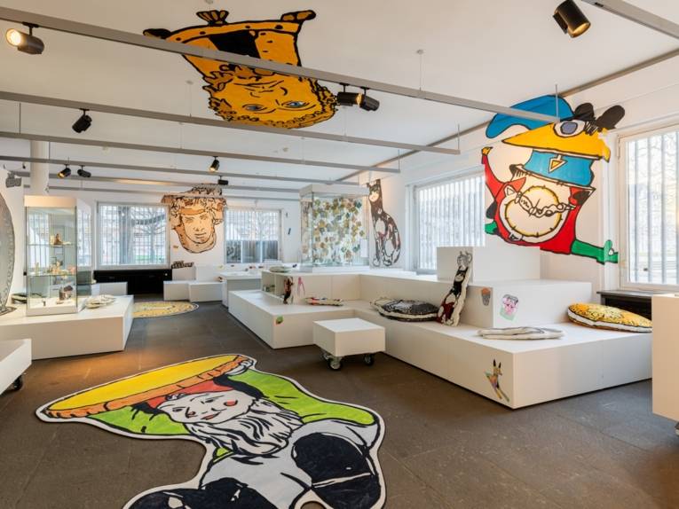 Blick in das Mitmachfoyer Sammler*s. Zu sehen sind großflächige bunte Paste-Ups an den Wänden und bunte Teppiche auf dem Boden, eine Vitrine mit Pixibüchern in der Bildmitte sowie eine Vitrine mit Objekten am linken Bildrand. Die weiße Sitzlandschaft bedecken teilweise bunte Kissen.