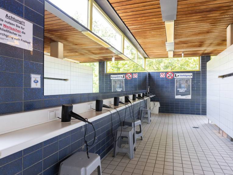 In einem Gebäude, alles gekachelt, links Spiegel und Föne, rechts Wand mit Kleiderhaken