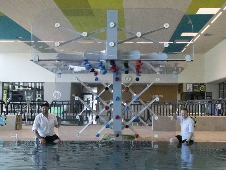 Zwei Männer, die am Beckenrand im Hallenbad sitzen, zwischen ihnen eine Kletterwand