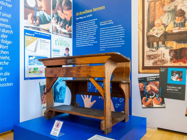 Alte Bank f&uuml;r Sch&uuml;ler in der Ausstellung, im Hintergrund Abbildungen und Texttafeln