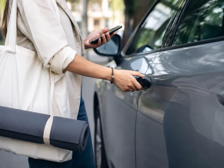 Una donna con una borsa e un tappetino da yoga a tracolla tiene il cellulare nella mano sinistra e con la destra apre la portiera di un’auto grigia.