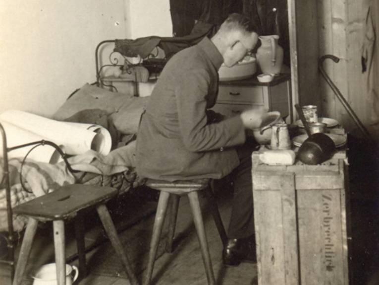 Mann essend auf einem Hocker vor einer Kiste sitzend, dahinter ein Bett, schwarz-weiß Aufnahme