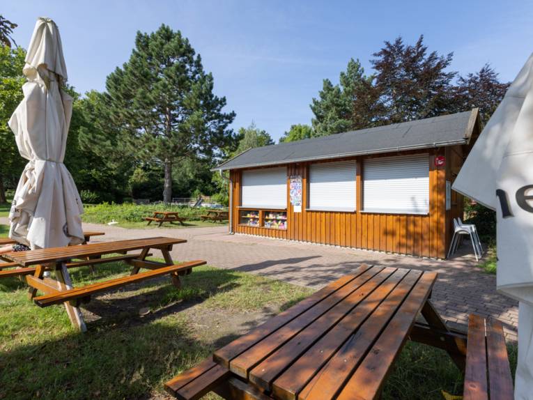 Holzbänke und Tische, zusammengeklappte Sonnenschirme, im Hintergrund ein Kiosk mit Holzwänden