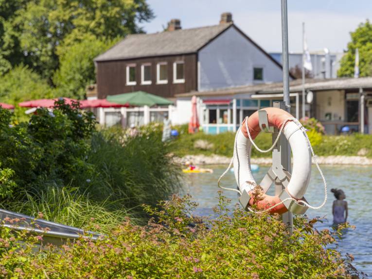 Flacher Badebereich mit Rettungsring im Vordergrund, im Hintergrund ein Geb&auml;ude