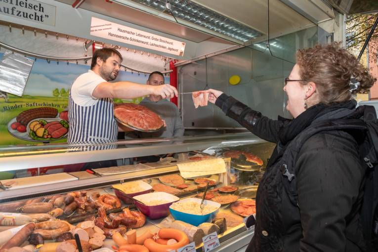 Ein H&auml;ndler bietet einer Kundin eine Scheibe Schinken zum probieren an.