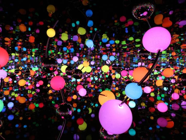 Zu sehen ist eine Lichtinstallation von Yayoi Kusama. Bunt leuchtende Kungeln in unterschiedlichen Farben sind in einem dunklen aber spiegelnden Raum verteilt.