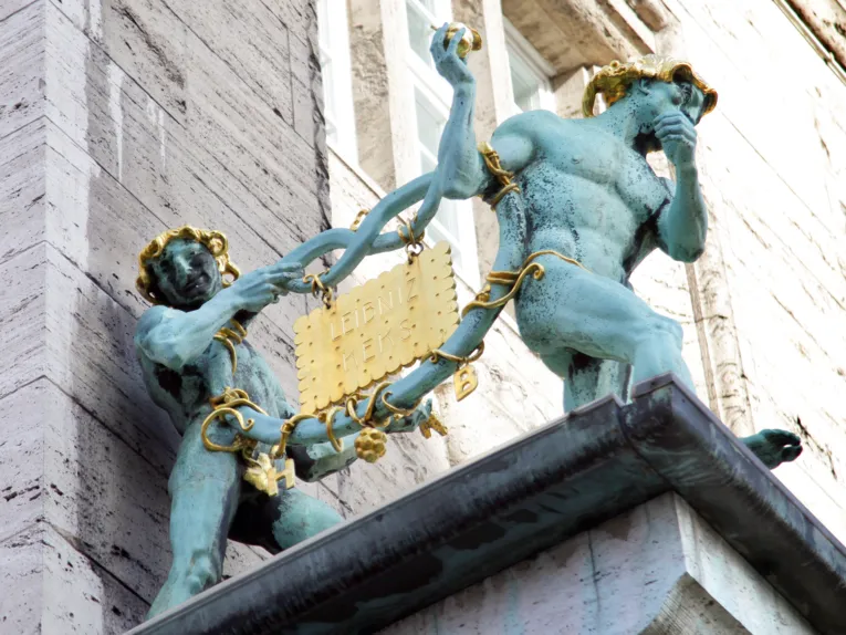 Zwei bronzene Figuren, die zusammen eine ebenfalls Bronzene Brezel tragen. In diese Brezel ist ein goldener Keks eingehängt - er hat die Aufschrift "Leibniz Keks".