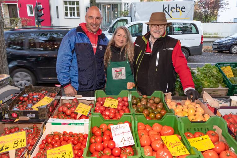 Verkaufspersonal steht l&auml;chelnd hinter einem Gem&uuml;sestand.