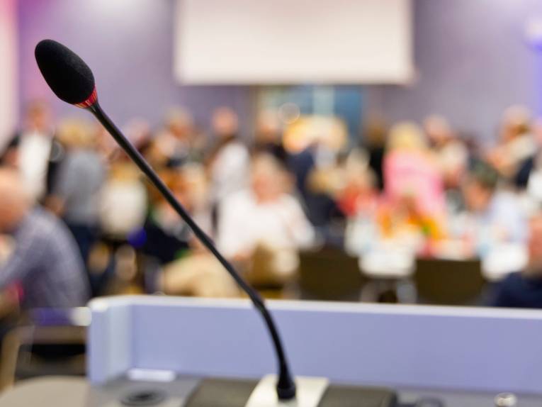 Im Vordergrund ist ein Mikrofon, festgemacht an einem Mikrofonständer, dahinter ist unscharf Publikum zu erkennen.