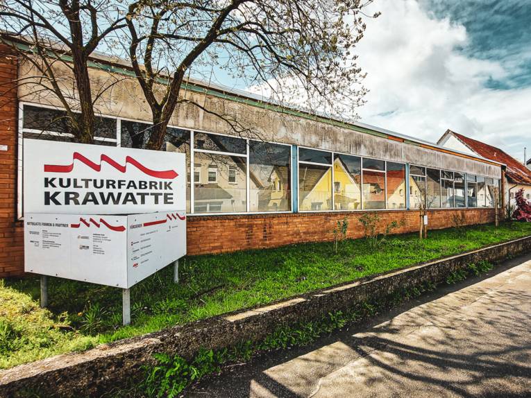 Vor einem langgezogenen Gebäude steht ein Schild, dort steht "Kulturfabrik Krawatte".