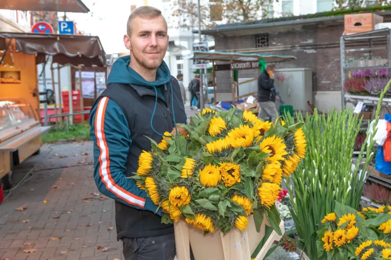 En mand holder en kasse fuld af gule blomster op mod kameraet.