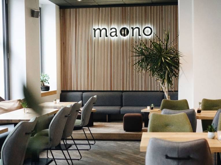 Restoran on sisustatud puidust laudadega, mille ääres seisavad hallid ja rohelised riidest toolid. Taustal on näha puidust seina, millel on restorani nimi „maono“.