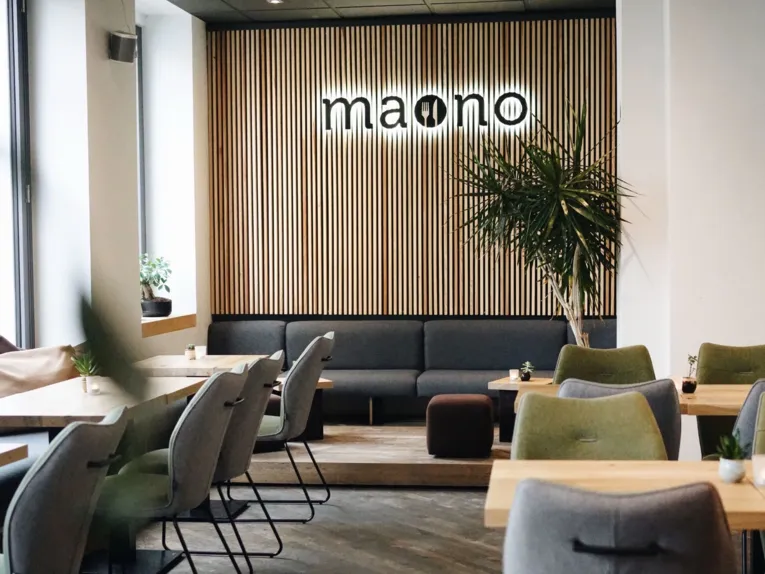 Ristorante arredato con tavoli in legno, attorno ai quali sono disposte sedie in tessuto grigio e verde. Sullo sfondo si vede una parete in legno con la scritta “maono”.