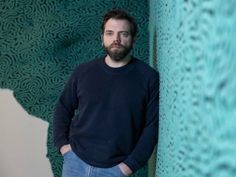 Zu sehen ist der Künstler Jérémy Gobé. Er steht an eine Wand gelehnt, die mit seiner "Korallenkunst" beklebt ist.