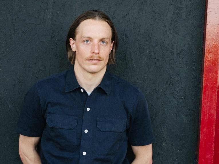 Zu sehen ist ein junger Mann in einem dunkelblauen, kurzärmeligen Hemd.