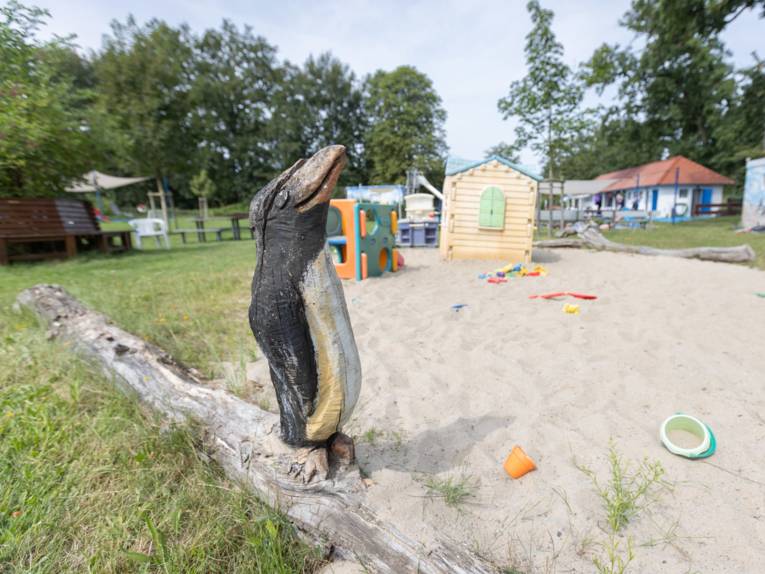 Im Vordergrund geschnitzter Pinguin, Sandkiste mit Spielhaus und Klettermöglichkeit