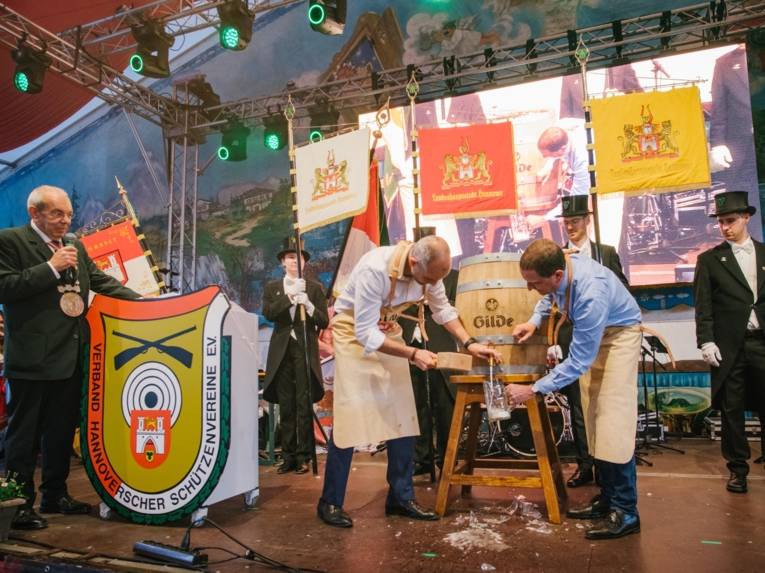 Mehrere Personen auf einer Bühne. Darauf steht ein Fass, das angeschlagen wird.