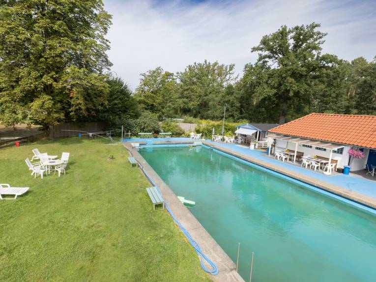 Schwimmbecken, im Hintergrund ein Gebäude und diverse Sitzmöglichkeiten sowohl vor dem Gebäude als auch auf der Wiese