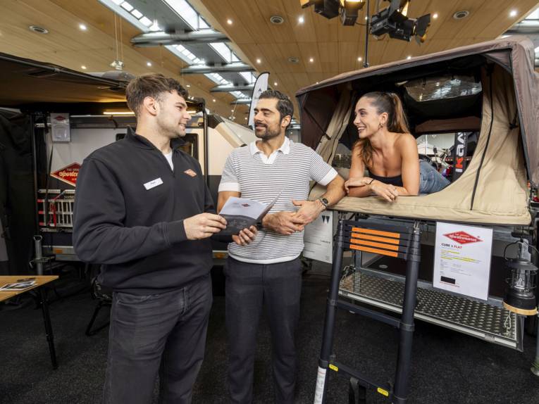 Eine Frau liegt in einem Dachzelt und zwei Männer stehen davor.