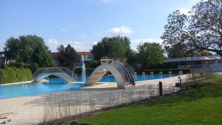 A piscina exterior cheia de água do complexo aquático coberto e ao ar livre de Burgdorf, sem banhistas, num dia de bom tempo, com um grande escorrega divertido, uma torre de saltos e uma ponte