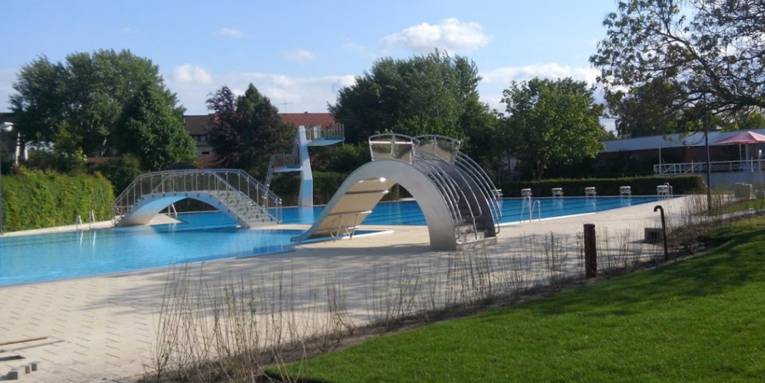 Das mit Wasser gefüllte Aussenbecken des Hallenfreibades Burgdorf ohne Badegäste bei schönem Wetter mit breiter Spaßrutsche, Sprungturm und Brücke