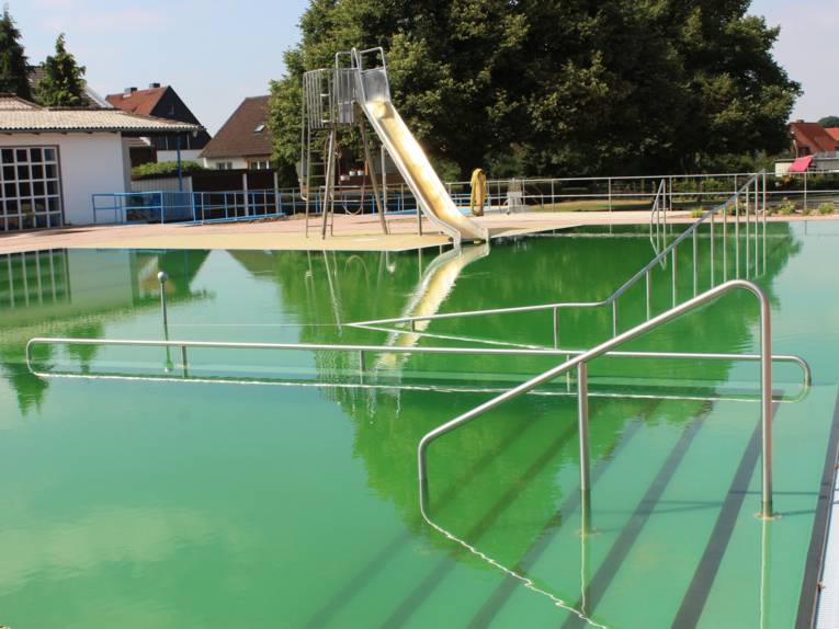 Ein Schwimmbecken, im Hintergrund eine Rutsche mit grünlich schimmerndem Wasser. Im Vordergrund Stufen, die ins Wasser führen.