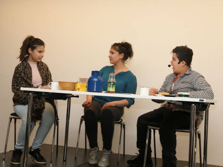 Zwei Schülerinnen und ein Schüler sitzen an einem Tisch auf einer kleinen Bühne.