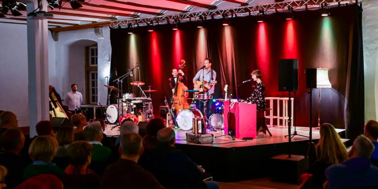 Saal im Schloss Landestrost mit Blick auf eine kleine beleuchtete Bühne und davor mehrere Stuhlreihen mit Publikum.