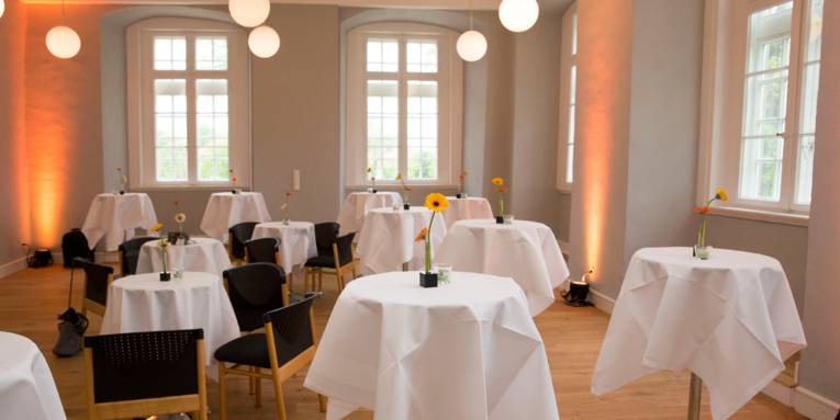 Ein Saal mit großen Fenstern, indem einige runde Stehtische sowie einige Tische mit Bestuhlung stehen. Auf allen Tischen ist eine weiße Tischdecke und einzelne Blumendekoration. Der raum wird durch mehrere kugelförmige Deckenlampen beleuchtet.