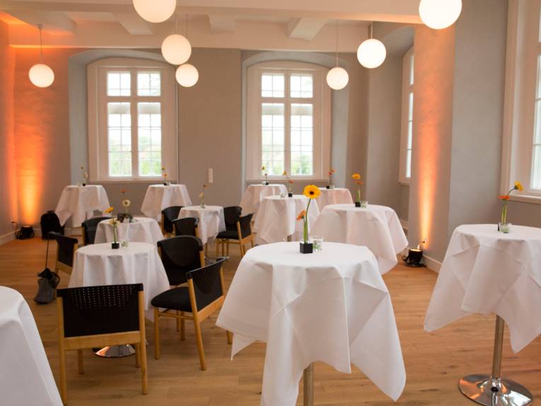 Ein Saal mit großen Fenstern, indem einige runde Stehtische sowie einige Tische mit Bestuhlung stehen. Auf allen Tischen ist eine weiße Tischdecke und einzelne Blumendekoration. Der raum wird durch mehrere kugelförmige Deckenlampen beleuchtet.