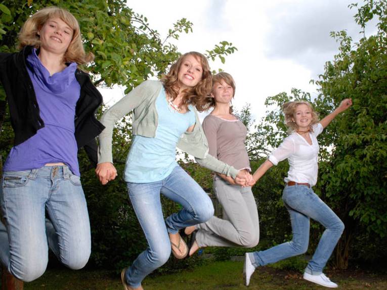 Vier strahlende junge Mädchen, die sich die Hände reichen und einen Luftsprung machen.