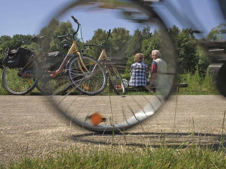 Durch die Speichen eines fahrenden Rades sieht man zwei Radfahrer, die am Wegrand auf einer Bank Pause machen