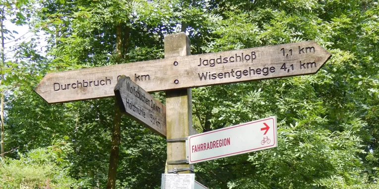 Et treskilt i skogen der blant annet "Durchbruch", "Jagdschloss" og "Wisentgehege" er merket med kilometerangivelser.