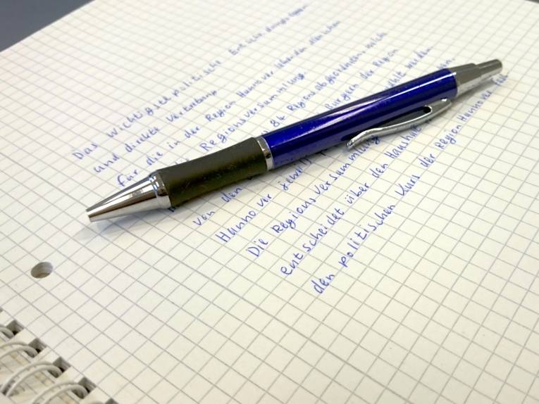 Das oberste Blatt eines Schreibblocks ist beschrieben. Obenauf liegt ein blauer Kugelschreiber.