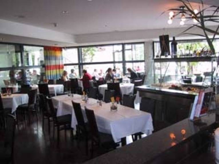 Im Restaurant stehen Tische mit weißen Decken und dunklen Stühlen darum. Im Hintergrund sitzen Gäste an einer Fensterfront.