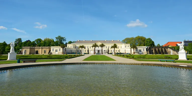 Elegantes Barockschloss mit symmetrischem Garten, Palmen und weißen Statuen, vor einem großen Wasserbecken unter blauem Himmel.