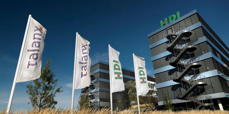 Vor einem modernen Bürogebäude sind Fahnen mit Firmenlogos gehisst.