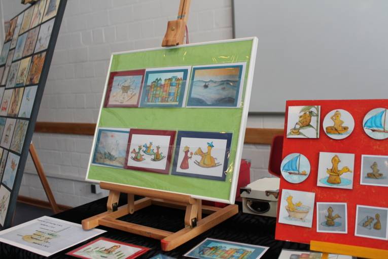 Auf einer Staffelei steht ein Bilderrahmen, auf dem verschiedene Postkarten befestigt sind. Auf den Karten sind kleine Figuren zu sehen.