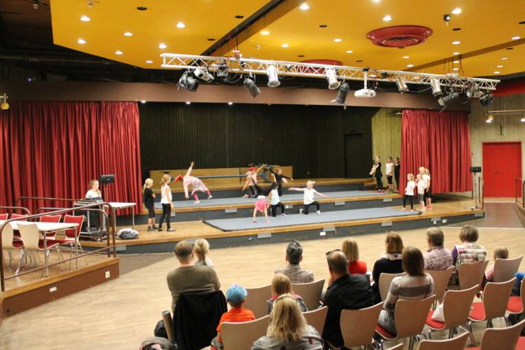 Man sieht Kinder, die auf einer Bühne akrobatische Kunststücke machen. Im Vordergrund sitzt das Publikum und schaut gebannt zu.