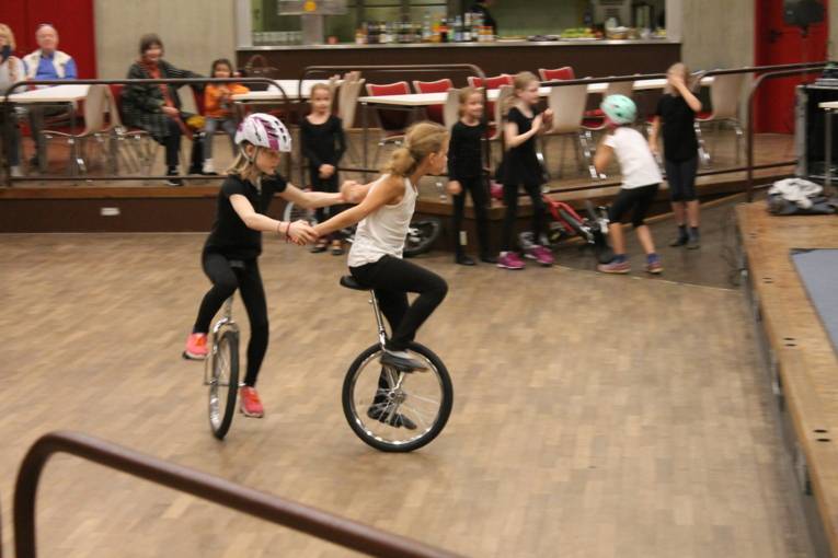 Zwei Mädchen halten sich beim Einradfahren an den Händen. Sie fahren gemeinsam einen Kreis.