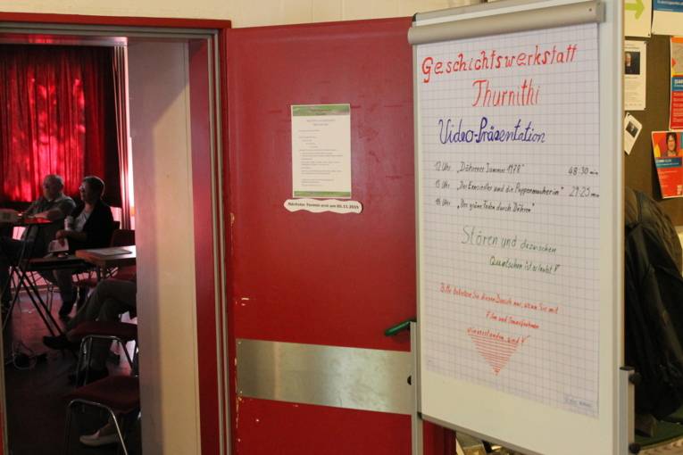 Auf dem Bild sieht man eine offene Tür. In dem Raum sitzen Menschen und schauen einen Film. Neben der Tür steht ein Schild mit der Aufschrift: Geschichtswerkstatt Thurnithi.