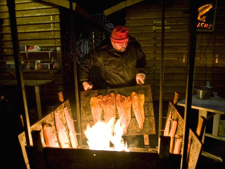 A burly man wearing a red cap is carrying a wooden frame with salmon fillets hanging from it to be smoked.
