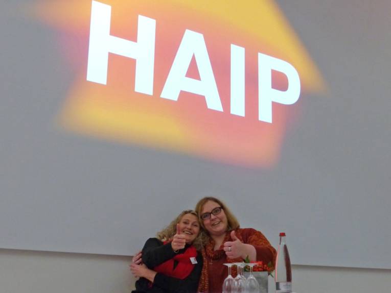 Zwei Frauen zeigen unter dem HAIP-Logo mit dem Daumen nach oben