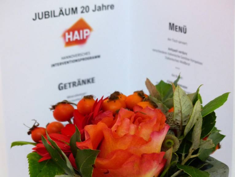 Ein Blumenstrauß mit Rosen und Hagebutten in den Farben des orange-roten HAIP-Logos vor dem aufgeklapptem Programmheft zum 20jährigen Bestehens der Initiative